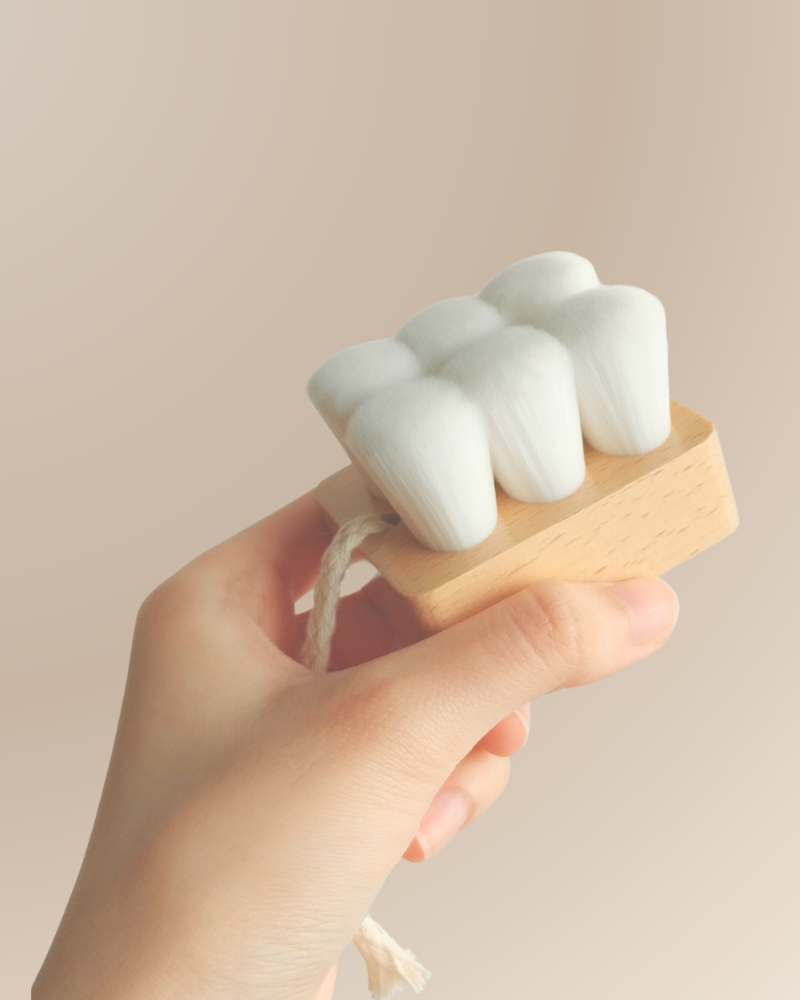 A hand holds the IREN Shizen Facial Cleansing Brush with ultra-soft, densely packed white bristles and a rope loop on the handle for gentle cleansing.