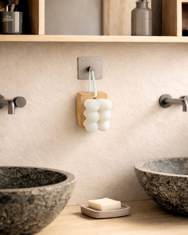 A modern bathroom with two stone sinks, wall-mounted faucets, a counter bar of soap, and the IREN Shizen Facial Cleansing Brush with ultra-soft bristles hanging on the wall between the sinks.