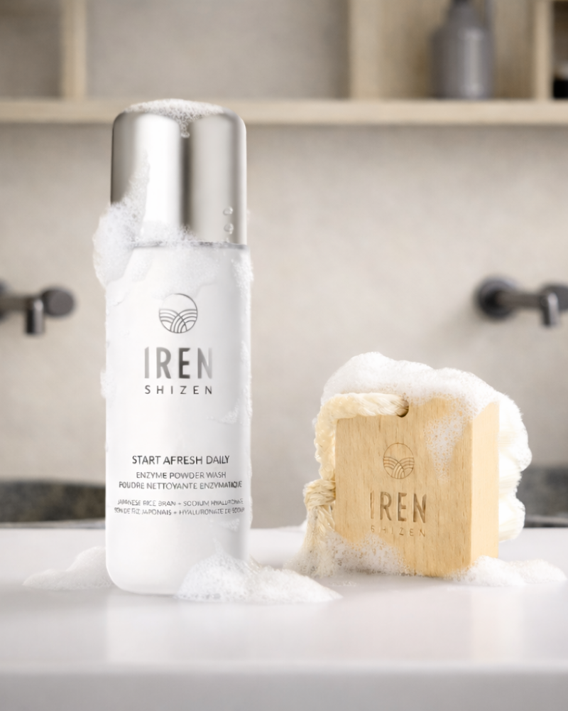 An IREN Shizen Start Afresh Daily Enzyme Powder Wash and an IREN Shizen Facial Cleansing Brush with ultra-soft bristles and foam rest on a bathroom counter, ready to offer a gentle cleanse.