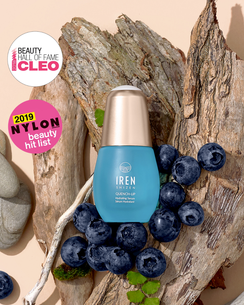 A blue QUENCH-UP Hydrating Serum bottle by IREN Shizen is surrounded by blueberries, driftwood, stones, and CLEO and NYLON award stickers—ideal for plump skin with Hyaluronic Acid.
