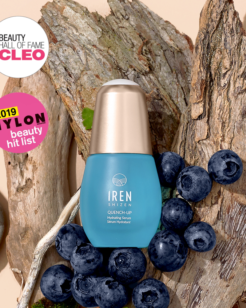 A blue bottle of IREN Shizen QUENCH-UP Hydrating Serum rests on tree bark with fresh blueberries. CLEO Beauty Hall of Fame and NYLON Beauty Hit List badges highlight its hyaluronic acid formula for plump, hydrated skin.