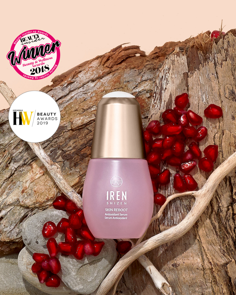 A bottle of IRÉN Shizen SKIN REBOOT Antioxidant Serum is shown on wood with pomegranate seeds, emphasizing its skin revitalization benefits and 2018/2019 beauty award wins.