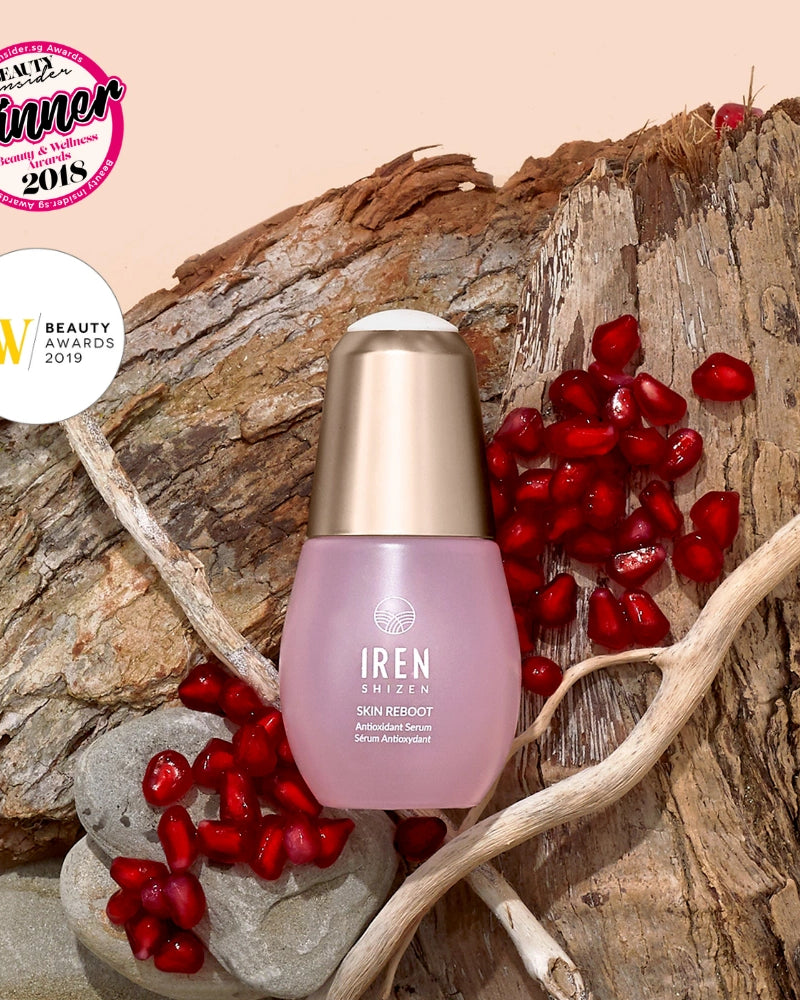 A bottle of IREN Shizen SKIN REBOOT Antioxidant Serum rests on wood with pomegranate seeds, rocks, and two beauty award badges, emphasizing its pomegranate extract for reducing dark circles.