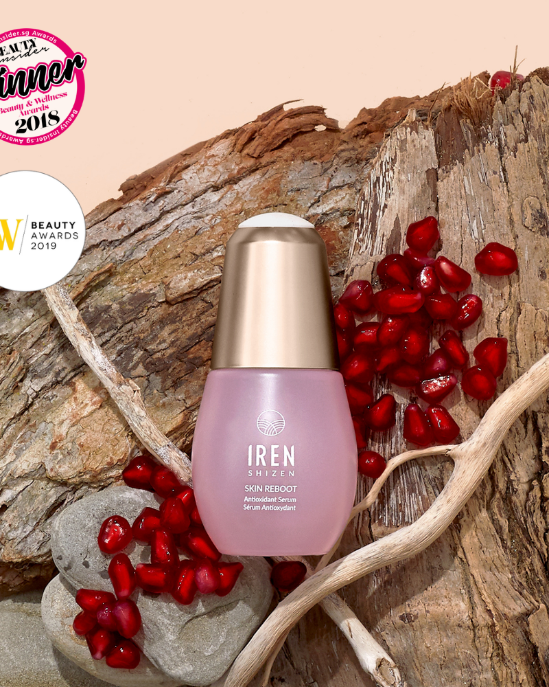 A bottle of IREN Shizen SKIN REBOOT Antioxidant Serum sits on tree bark among pomegranate seeds and twigs, with two beauty award badges displayed in the top left corner.