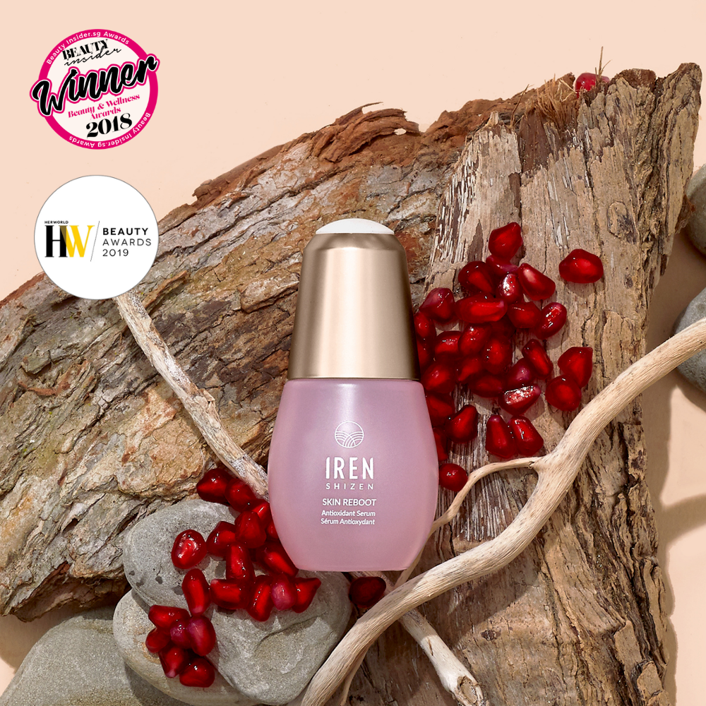 A bottle of IREN Shizen SKIN REBOOT Antioxidant Serum sits on tree bark among pomegranate seeds and twigs, with two beauty award badges displayed in the top left corner.