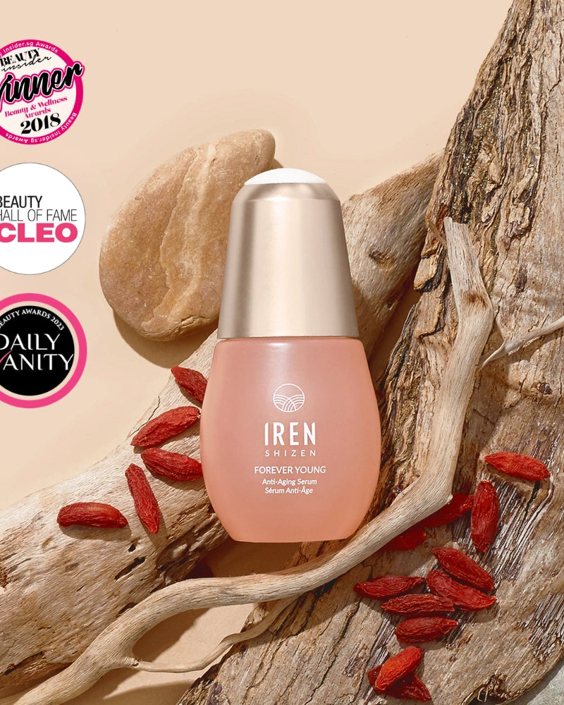 A bottle of IREN Shizen FOREVER YOUNG Anti-Aging Serum rests on driftwood with goji berries, framed by three award badges in the top left, emphasizing its anti-aging properties and collagen support.