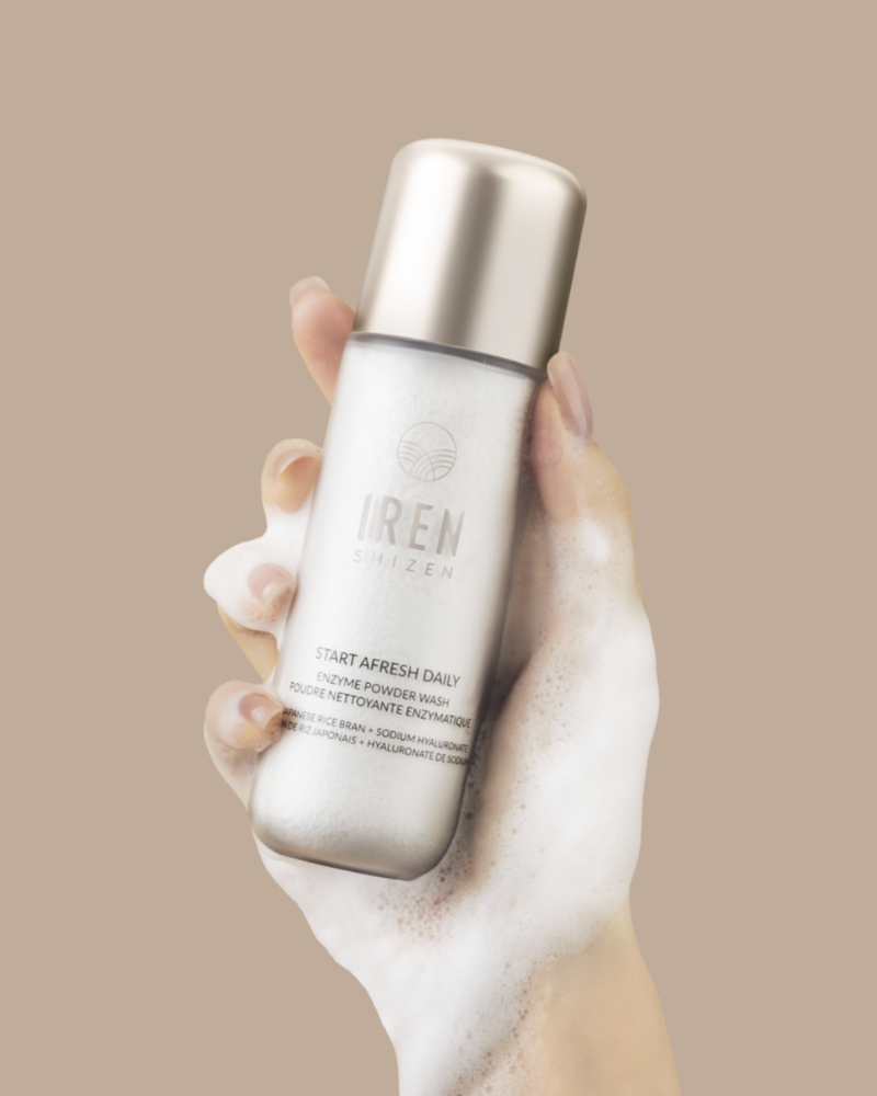 A hand covered in foam holds a bottle of Fast Bundle Cloud Cleansing Kit, a gentle enzyme cleanser, against a beige background.