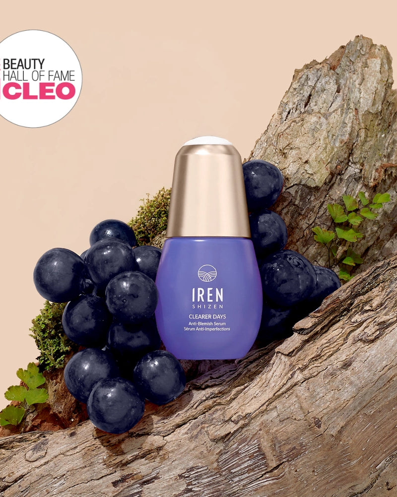 A bottle of IREN Shizen CLEARER DAYS Anti-Blemish Serum sits on a tree trunk with grapes and ferns, featuring a "Beauty Hall of Fame CLEO" award badge in the top left corner.