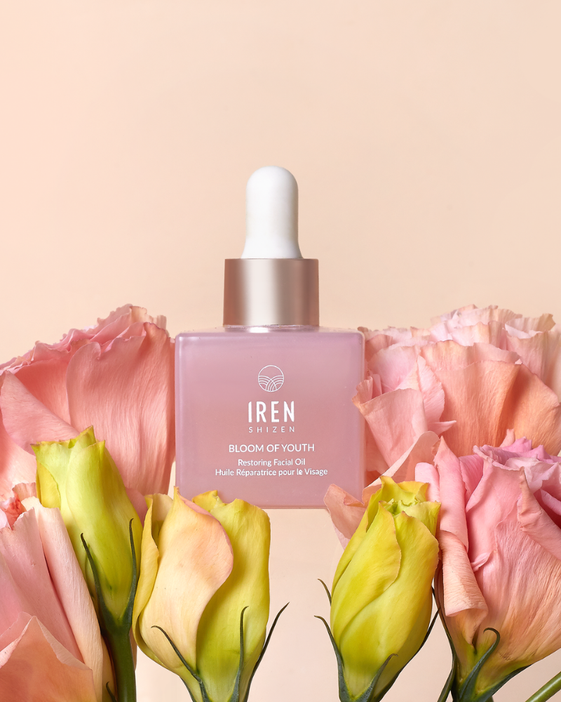 A pink dropper bottle labeled "BLOOM OF YOUTH Restoring Facial Oil" by IREN Shizen sits among pastel pink and yellow flowers, emphasizing its lightweight formula for skin barrier repair against a light peach background.