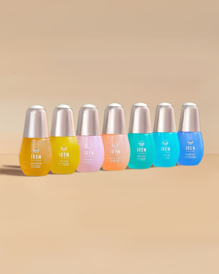 Seven colorful bottles of IREN Shizen’s MOISTURE SANDWICH Skincare Kit with silver caps are arranged in a row against a neutral background, designed to boost hydration and enhance serum absorption for radiant skin.