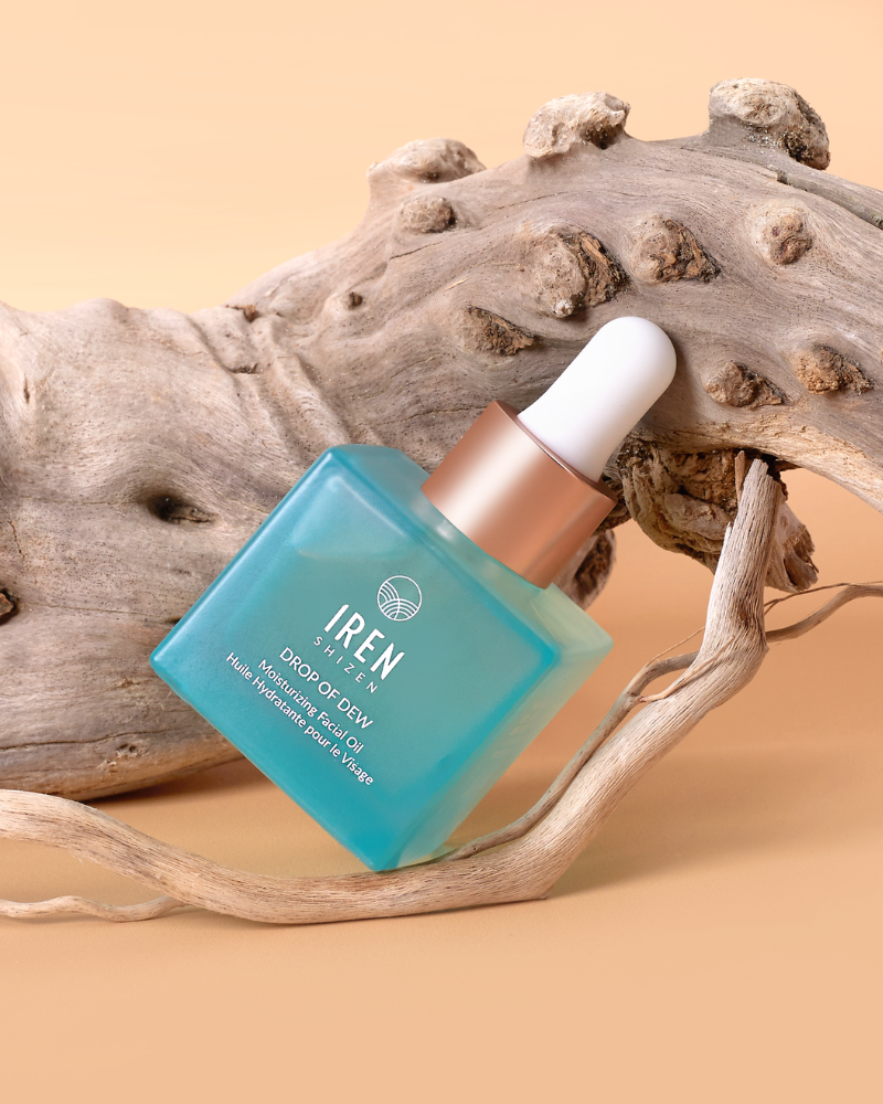 A blue glass dropper bottle labeled "IREN Shizen DROP OF DEW Moisturizing Facial Oil," a multi-tasking oil with cold-pressed oils, sits against a curved piece of driftwood on a beige background.