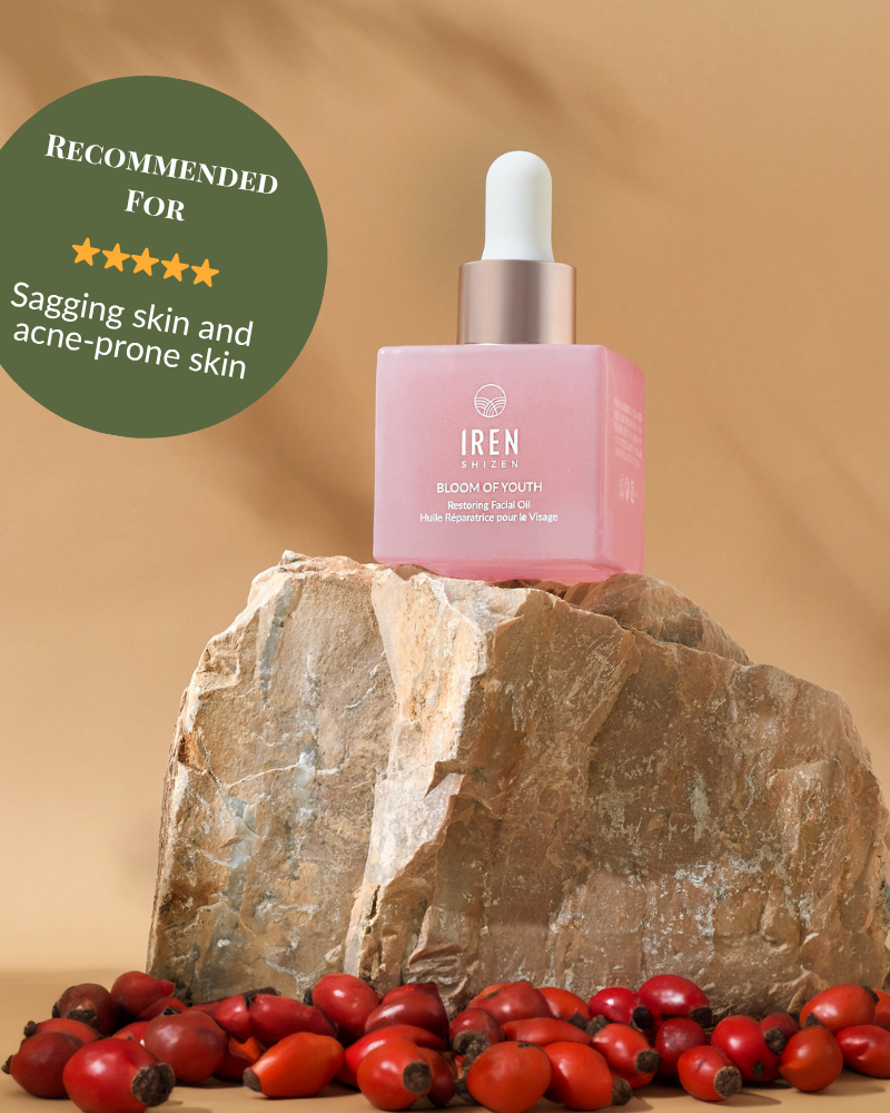 A pink bottle of BLOOM OF YOUTH Restoring Facial Oil by IREN Shizen with a dropper sits on a rock among rosehips; its green label highlights prebiotics and postbiotics for supporting the skin barrier, targeting sagging and acne-prone skin. #show-lang-en