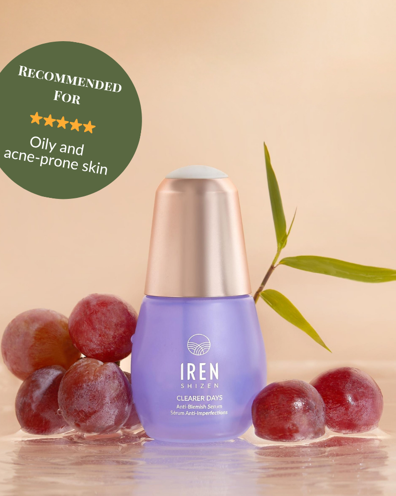 A bottle of IREN Shizen CLEARER DAYS Anti-Blemish Serum sits on a wet surface with red grapes, a green stem, and a badge recommending it for sebum balance and oily, acne-prone skin. #show-lang-en