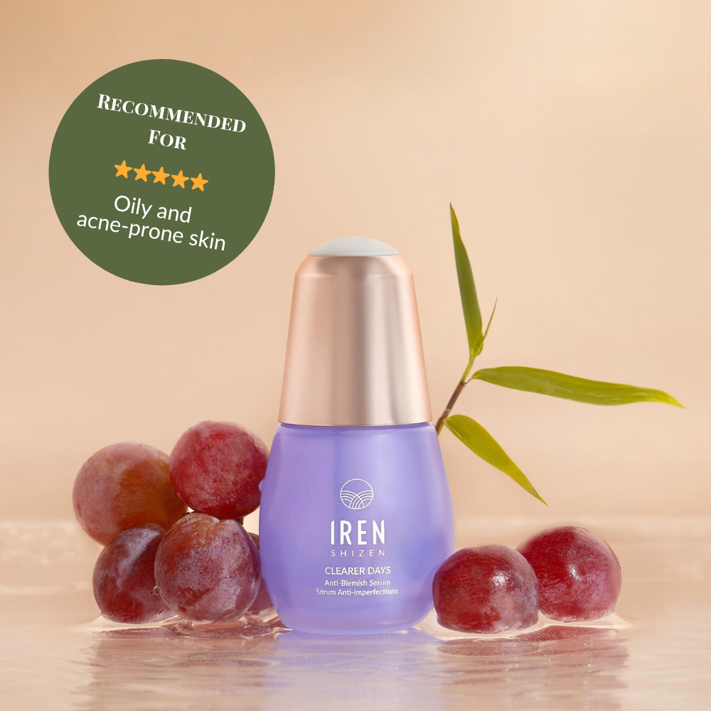 A bottle of IREN Shizen CLEARER DAYS Anti-Blemish Serum sits on a wet surface with red grapes, a green stem, and a badge recommending it for sebum balance and oily, acne-prone skin. #show-lang-en