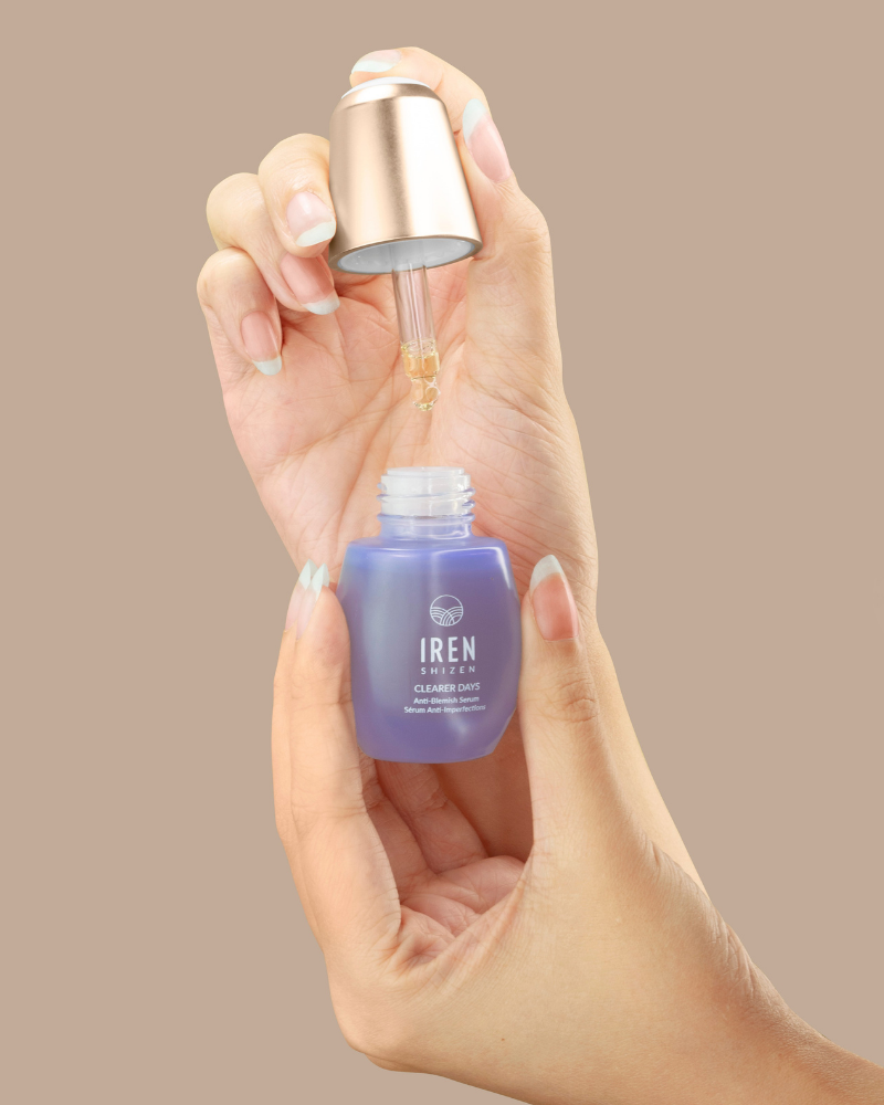 A hand holds a purple bottle of IREN Shizen CLEARER DAYS Anti-Blemish Serum with a dropper, dispensing liquid for sebum balance, against a plain beige background.
