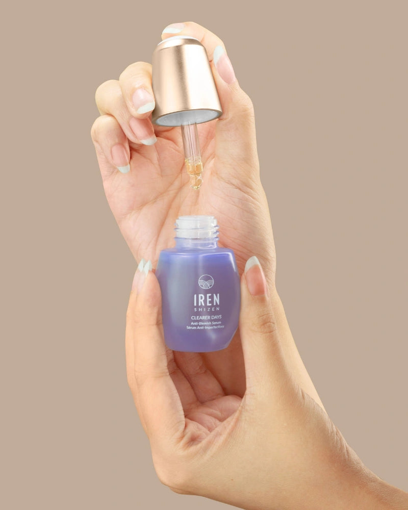 A hand holds a purple IREN Shizen CLEARER DAYS Anti-Blemish Serum bottle while another dispenses the serum with a dropper against a beige background, highlighting its acne treatment and sebum-balancing benefits.
