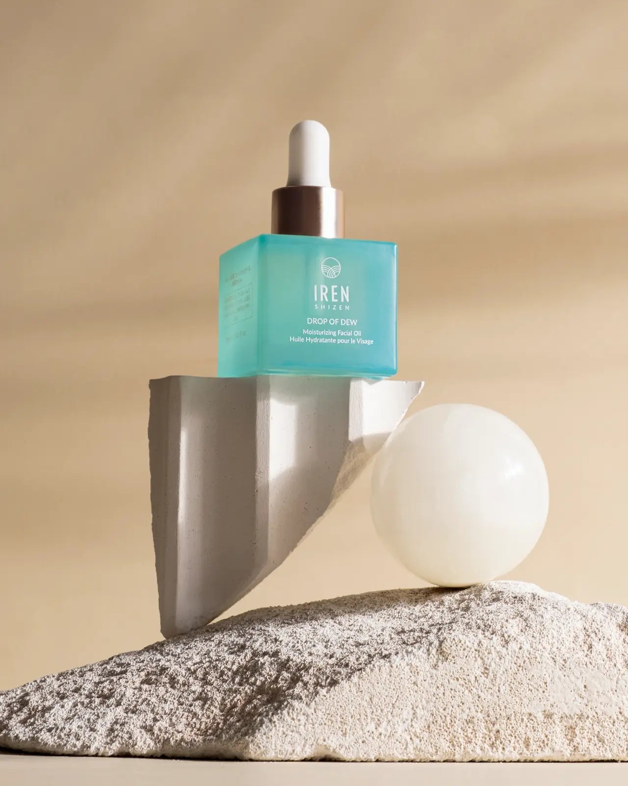 A blue GLOW ON THE GO Travel Light Kit by IREN Shizen balances on a broken stone slab with a white sphere and textured base, capturing travel elegance against a soothing beige background.