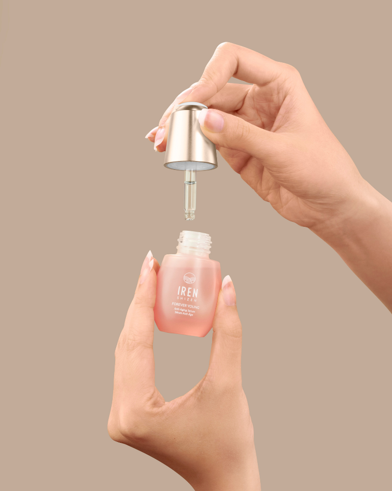A person holds a small bottle of IREN Shizen FOREVER YOUNG Anti-Aging Serum in one hand and removes the dropper cap with the other, showcasing its anti-aging benefits against a plain beige background.