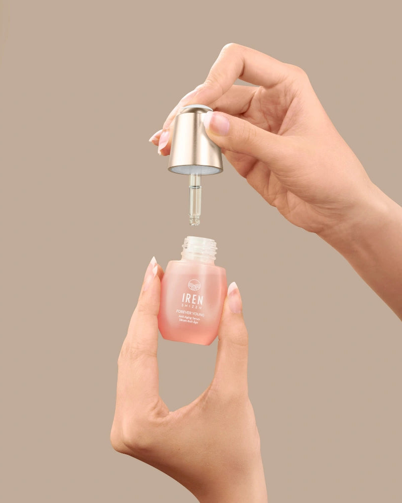 A person holds a dropper and a small glass bottle of pink FOREVER YOUNG Anti-Aging Serum by IREN Shizen, designed to support collagen production, against a plain beige background.