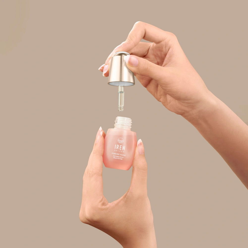 A person holds a dropper and a small glass bottle of pink FOREVER YOUNG Anti-Aging Serum by IREN Shizen, designed to support collagen production, against a plain beige background. #all-groups