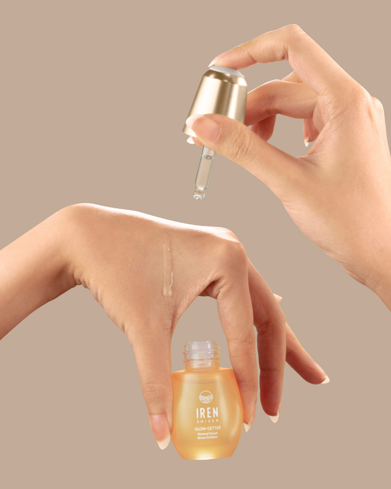 A hand applies a drop of IREN Shizen GLOW-GETTER Renewal Serum onto the back of another hand with a dropper against a beige background.