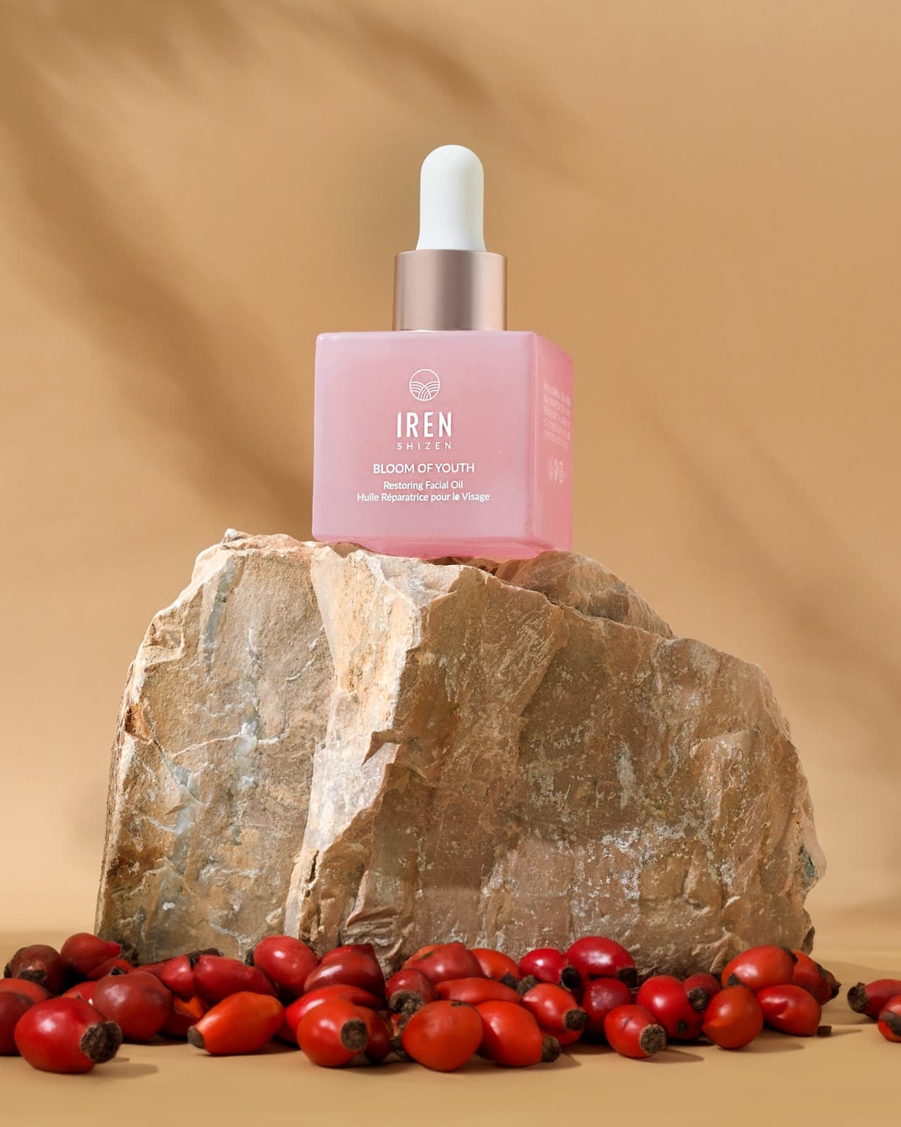 A pink dropper bottle labeled "IREN Shizen" from the SKIN GENIE PRO Hydration Set sits on a large rock, surrounded by rose hips and a Skin Genie Pro facial cleansing device, set against a beige background.