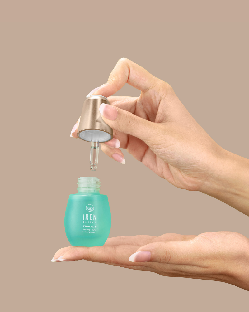 A hand dispenses KEEP CALM Soothing Serum by IREN Shizen from a blue bottle onto the palm, ideal for sensitive skin, with a plain beige background.