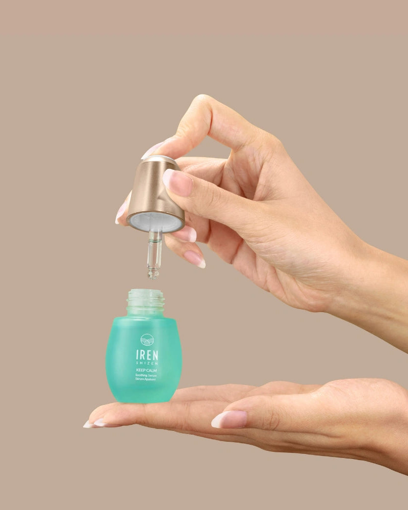 A person dispenses KEEP CALM Soothing Serum by IREN Shizen from a dropper bottle onto their palm against a plain beige background.