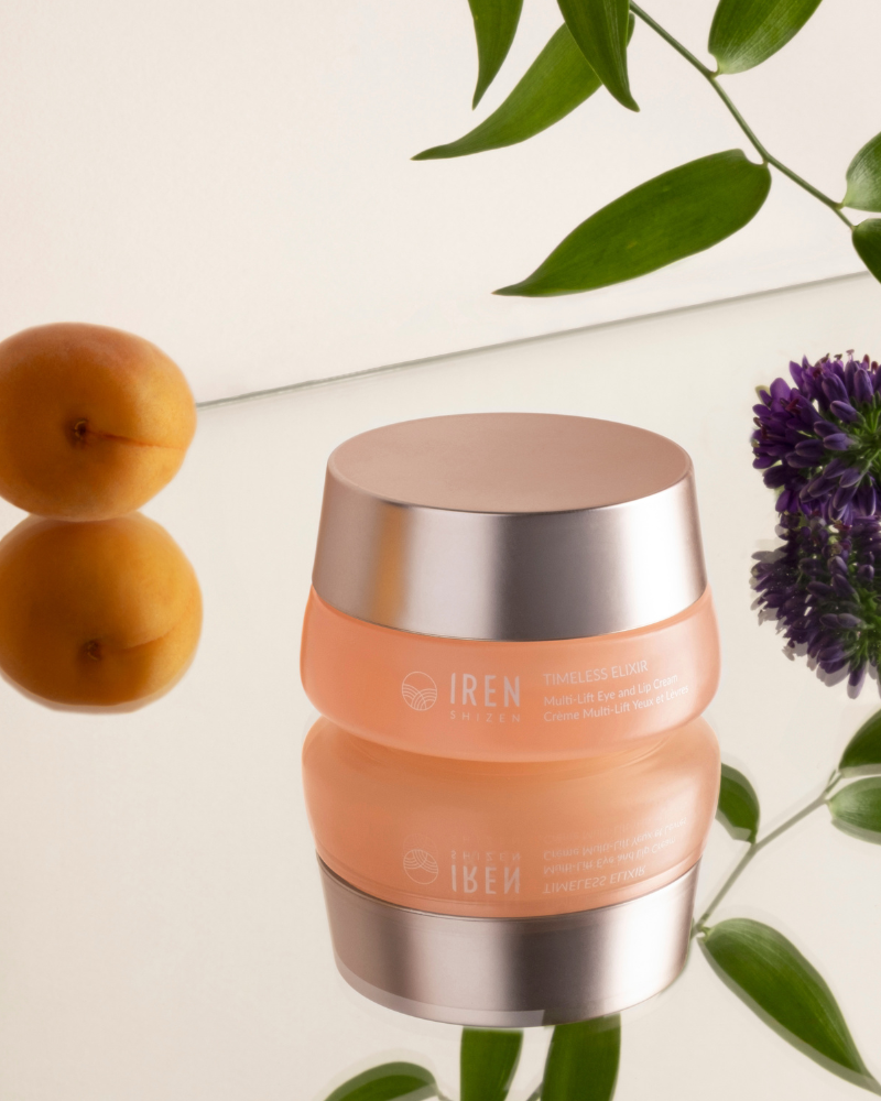 A jar of IREN Shizen TIMELESS ELIXIR Multi-Lift Eye and Lip Cream, celebrated for its anti-aging properties, is elegantly placed on a reflective surface surrounded by apricots, green leaves, and a purple flower.