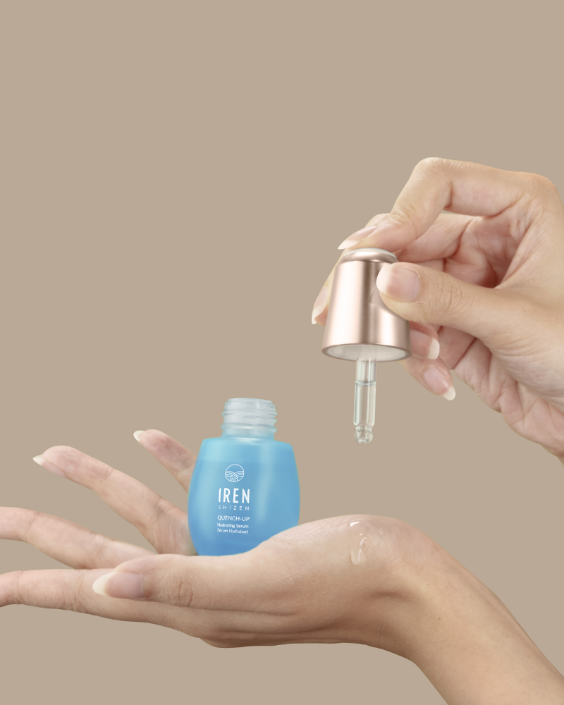 A hand holds a dropper above an open blue bottle of IREN Shizen QUENCH-UP Hydrating Serum, dispensing hyaluronic acid-infused liquid onto the palm against a beige background for plumper skin.