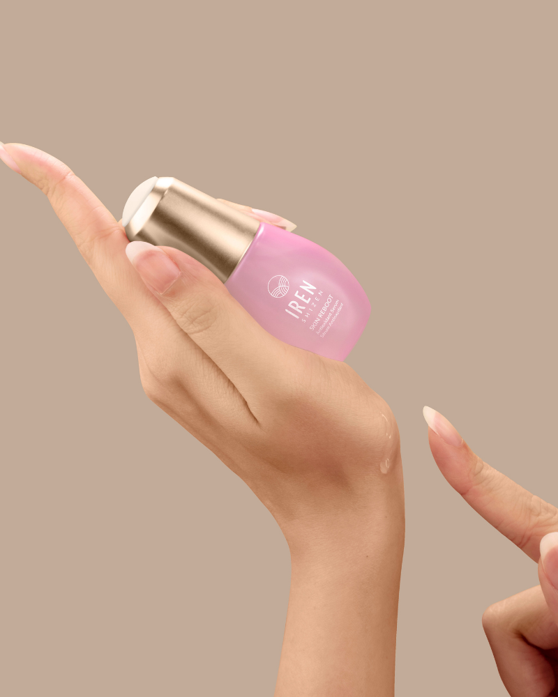 A person dispenses IREN Shizen’s SKIN REBOOT Antioxidant Serum, a pink formula, onto their fingertip from a small bottle against a plain beige background.