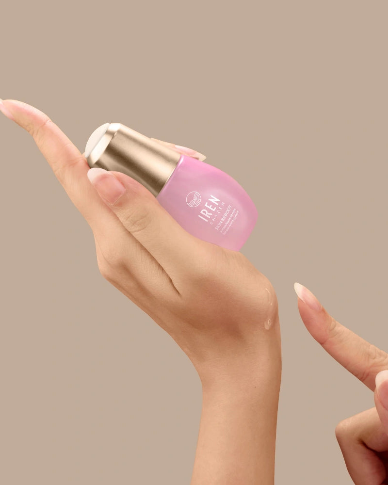 A hand holds a small pink bottle of SKIN REBOOT Antioxidant Serum by IREN Shizen with a gold cap, dispensing a drop onto the back of the hand. The serum features pomegranate extract to help reduce dark circles against a beige background.