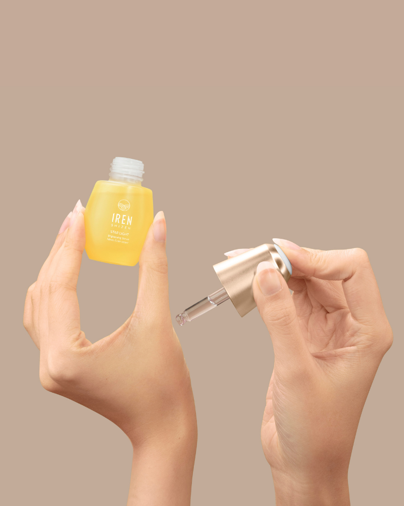 A person holds a small yellow bottle of IREN Shizen STAR LIGHT Brightening Serum in one hand and a dropper in the other, ready to target hyperpigmentation, against a plain beige background.