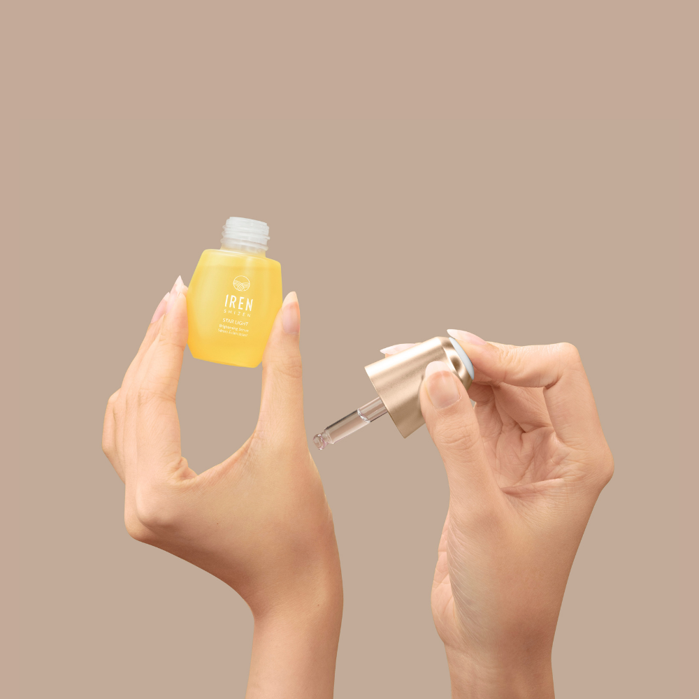 A person holds a small yellow bottle of IREN Shizen STAR LIGHT Brightening Serum in one hand and a dropper in the other, ready to target hyperpigmentation, against a plain beige background.