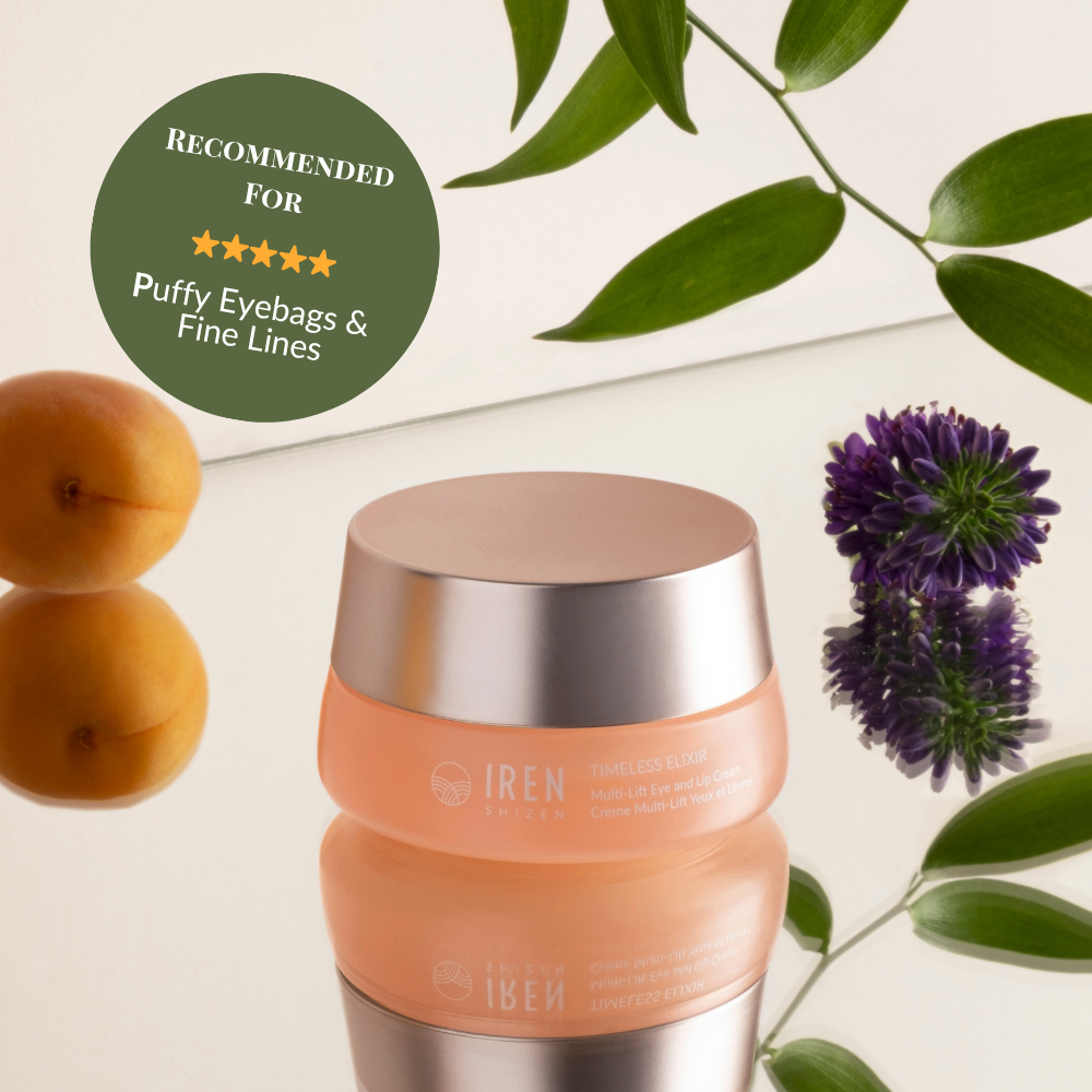 A peach IREN Shizen TIMELESS ELIXIR Multi-Lift Eye and Lip Cream jar sits on a reflective surface with leaves, purple flowers, and apricots. A green badge recommends it for treating puffy eyebags, fine lines, and dark circles. #show-lang-en