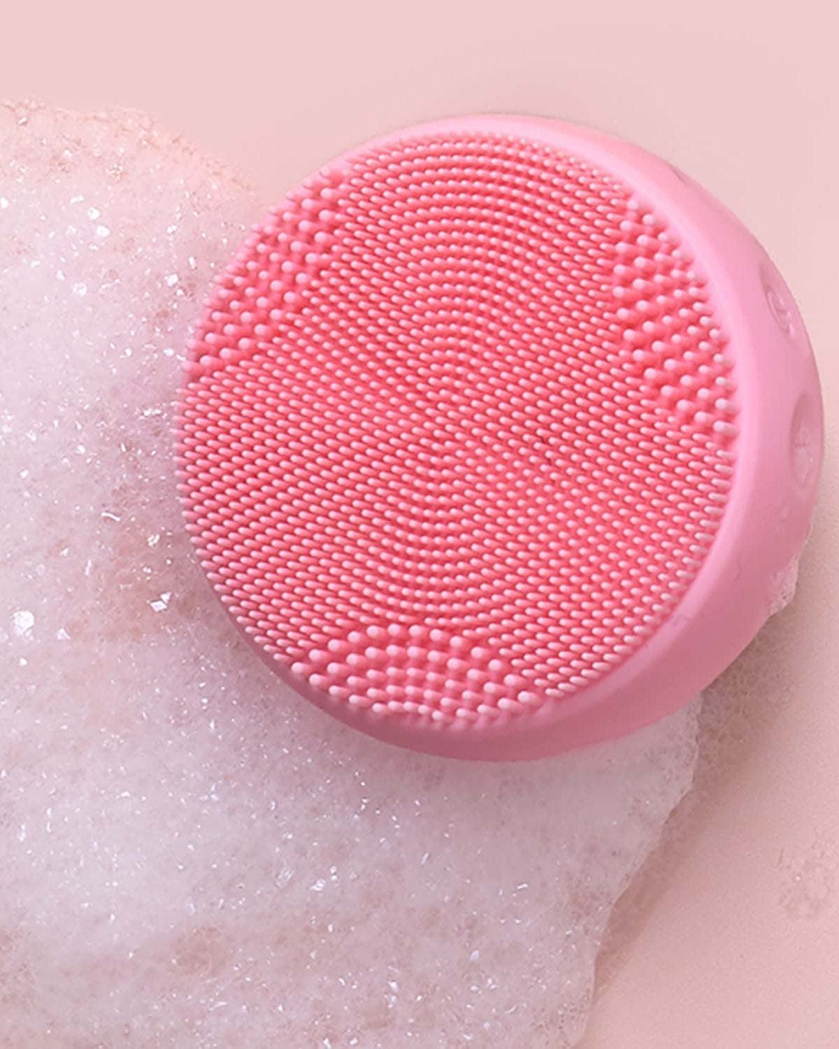 A round pink SKIN GENIE PRO Hydration Set facial cleansing device by IREN Shizen with textured bristles sits on a foamy surface against a light pink background.