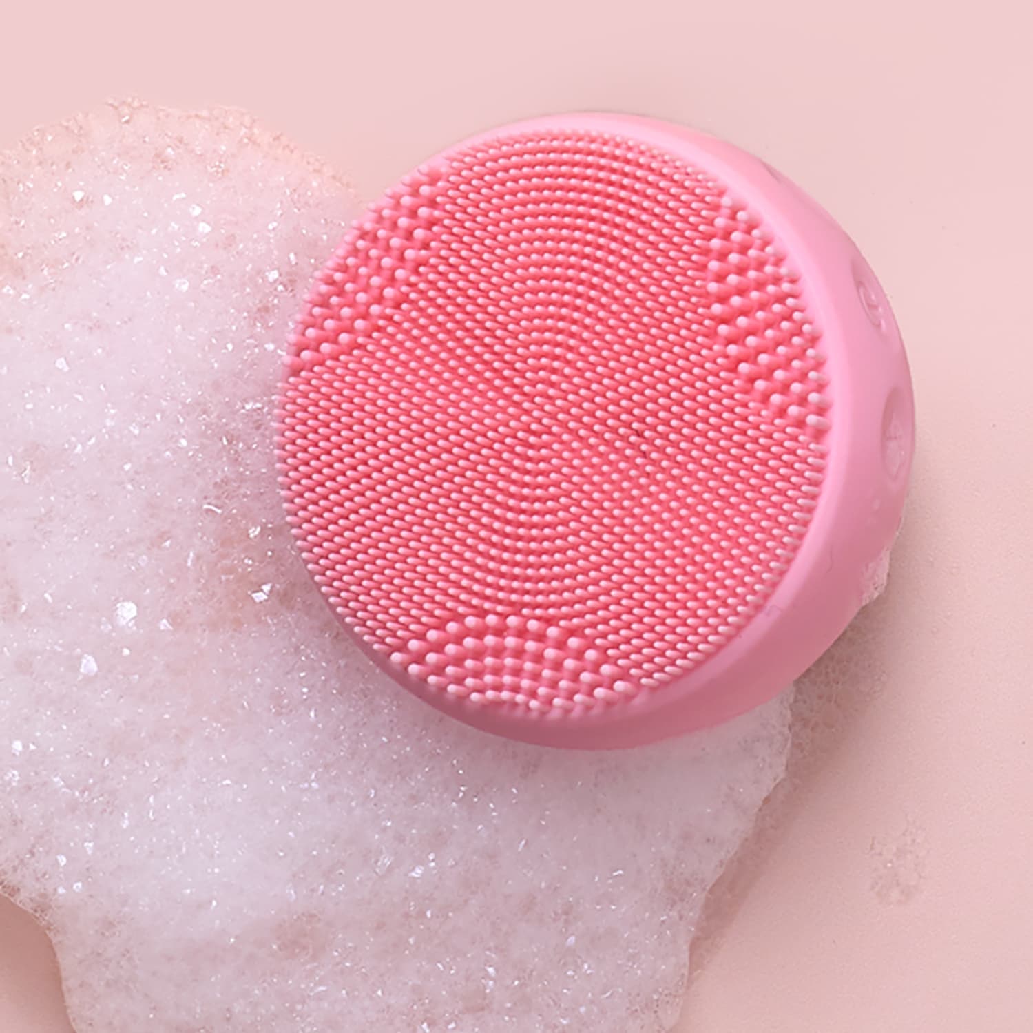 A round pink SKIN GENIE PRO Hydration Set facial cleansing device by IREN Shizen with textured bristles sits on a foamy surface against a light pink background.