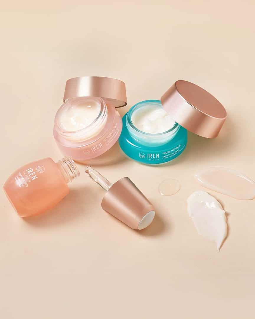 Three open jars of skincare cream—one blue and two pink—alongside a dropper bottle and cream samples on a beige surface, make up our MOISTURE SANDWICH Skincare Kit featuring Hydra Firming Cream and Antioxidant Water Cream by IREN Shizen.