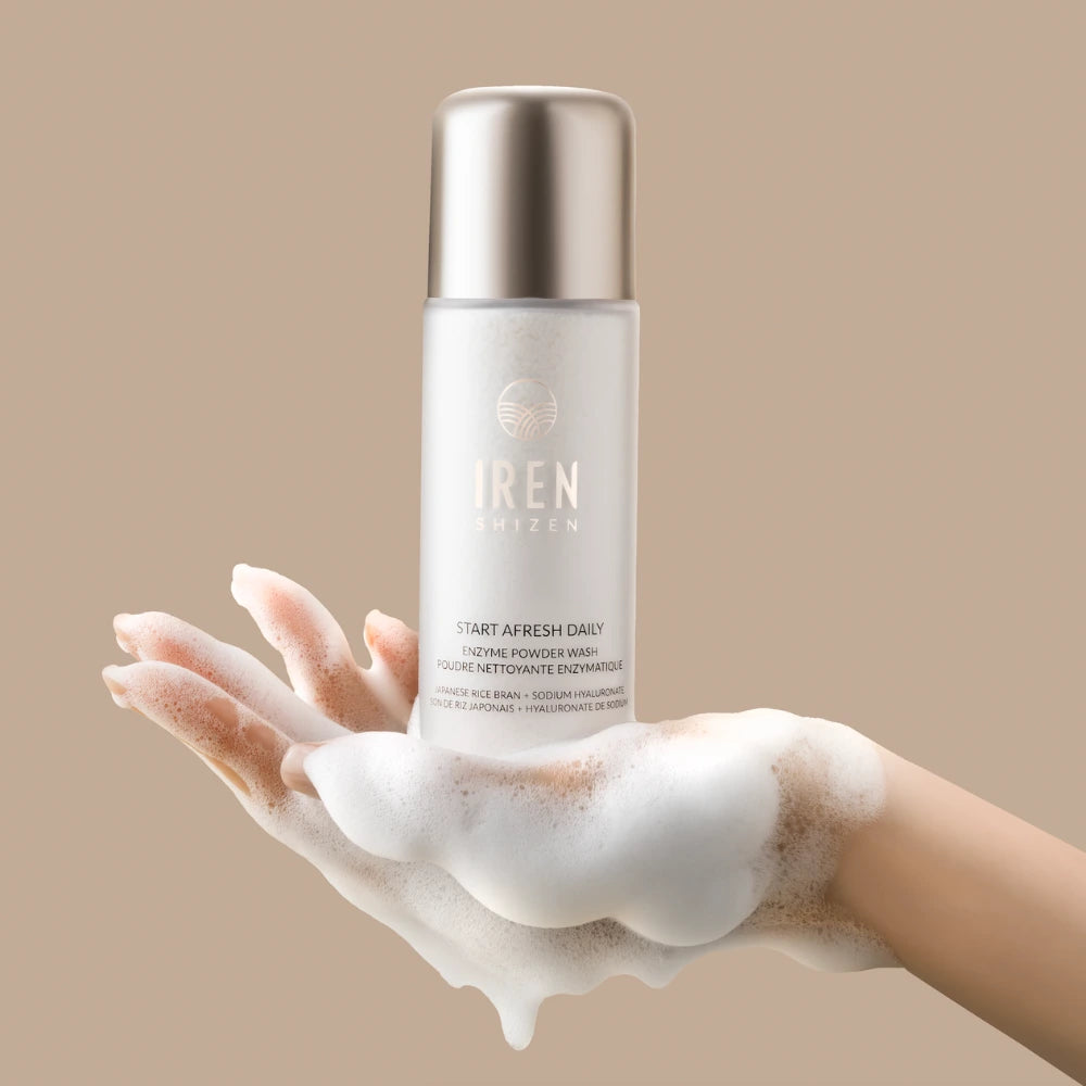 A foam-covered hand holds a bottle of IREN Shizen START AFRESH DAILY Enzyme Powder Wash—a gentle enzyme cleanser and daily exfoliant—against a beige background.