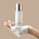 A foam-covered hand holds a bottle of IREN Shizen START AFRESH DAILY Enzyme Powder Wash—a gentle enzyme cleanser and daily exfoliant—against a beige background.