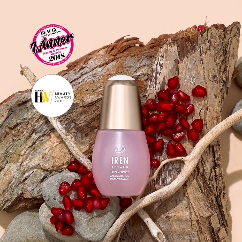 A bottle of IRÉN Shizen SKIN REBOOT Antioxidant Serum is shown on wood with pomegranate seeds, emphasizing its skin revitalization benefits and 2018/2019 beauty award wins.