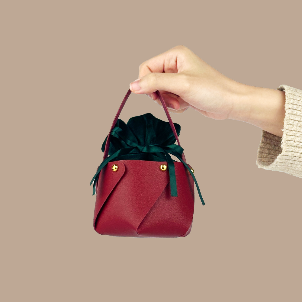 A hand holding the IREN Shizen FOR THE PALS Xmas Apple Gift Bag, a small red leather bag with a green drawstring pouch inside, against a neutral background.