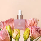 A pink dropper bottle labeled "BLOOM OF YOUTH Restoring Facial Oil" by IREN Shizen sits among pink and yellow flowers against a light beige background, highlighting its lightweight formula that supports skin barrier repair.
