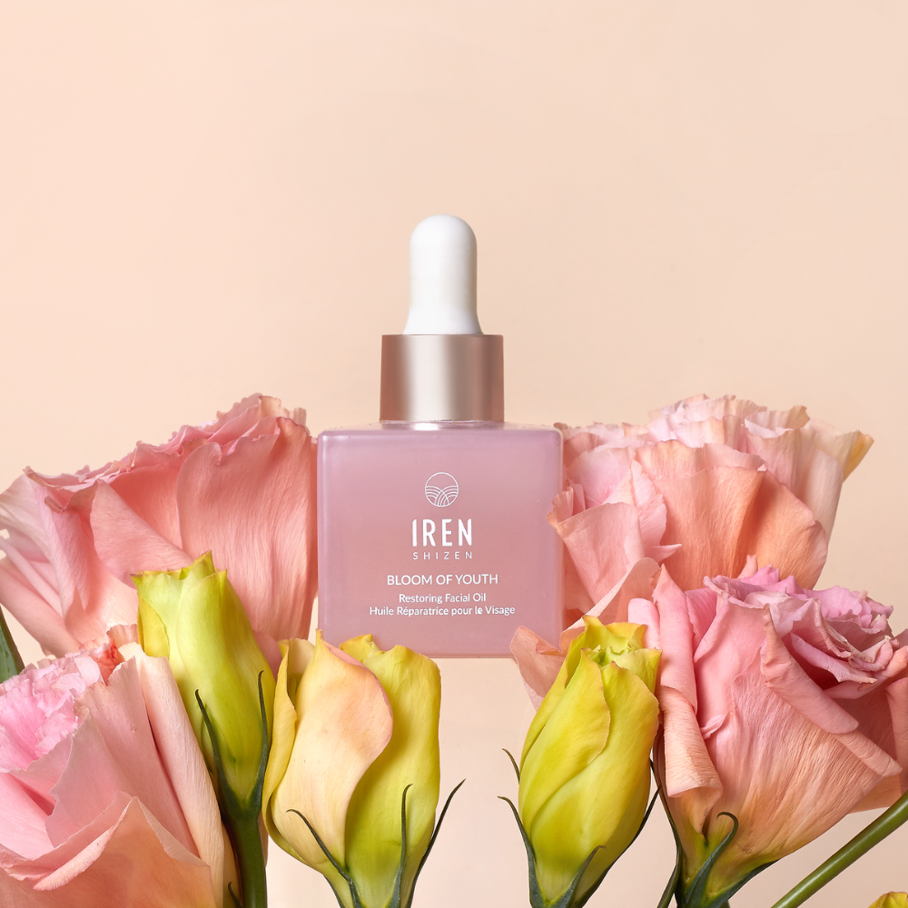 A pink dropper bottle labeled "BLOOM OF YOUTH Restoring Facial Oil" by IREN Shizen sits among pastel pink and yellow flowers, emphasizing its lightweight formula for skin barrier repair against a light peach background.