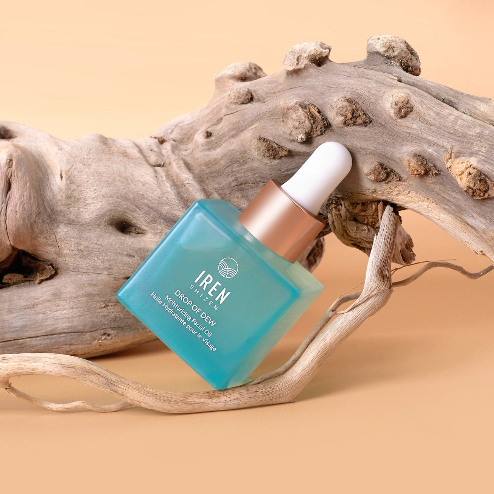 A blue dropper bottle labeled "IREN Shizen" containing DROP OF DEW Moisturizing Facial Oil for skin barrier repair rests on textured driftwood against a beige background.