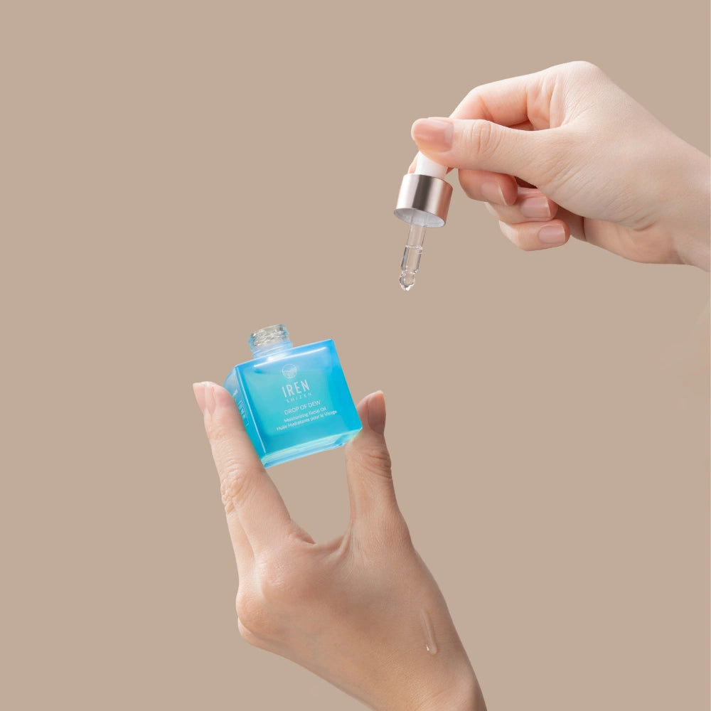 A hand holds IREN Shizen DROP OF DEW Moisturizing Facial Oil, dispensing the multi-tasking, cold-pressed oil for skin barrier repair with a dropper, set against a beige background.