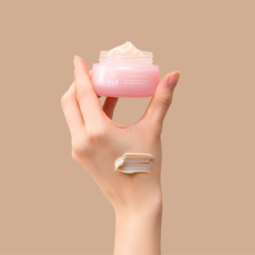 A hand holds an open jar of IREN Shizen FLOWERS IN BLOOM Antioxidant Water Cream against a beige background, with a swipe of the hydrating formula applied to the back of the hand.