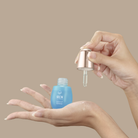 A hand holds a dropper above an open blue bottle of IREN Shizen QUENCH-UP Hydrating Serum, dispensing hyaluronic acid-infused liquid onto the palm against a beige background for plumper skin.