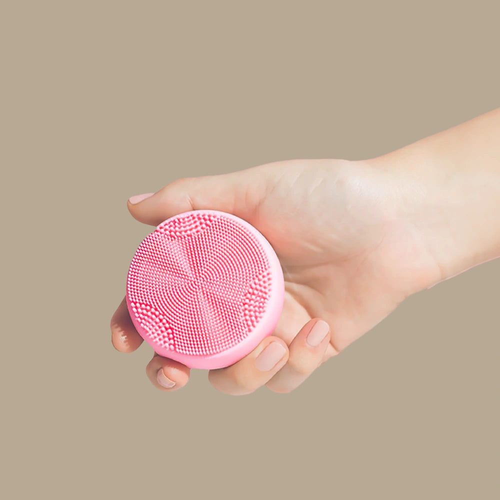 A hand holds the SKIN GENIE PRO Cleansing Brush + LED Light Therapy by IREN Shizen, featuring anti-bacterial silicone bristles, against a plain beige background.