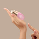A hand holds a small pink bottle of SKIN REBOOT Antioxidant Serum by IREN Shizen with a gold cap, dispensing a drop onto the back of the hand. The serum features pomegranate extract to help reduce dark circles against a beige background.